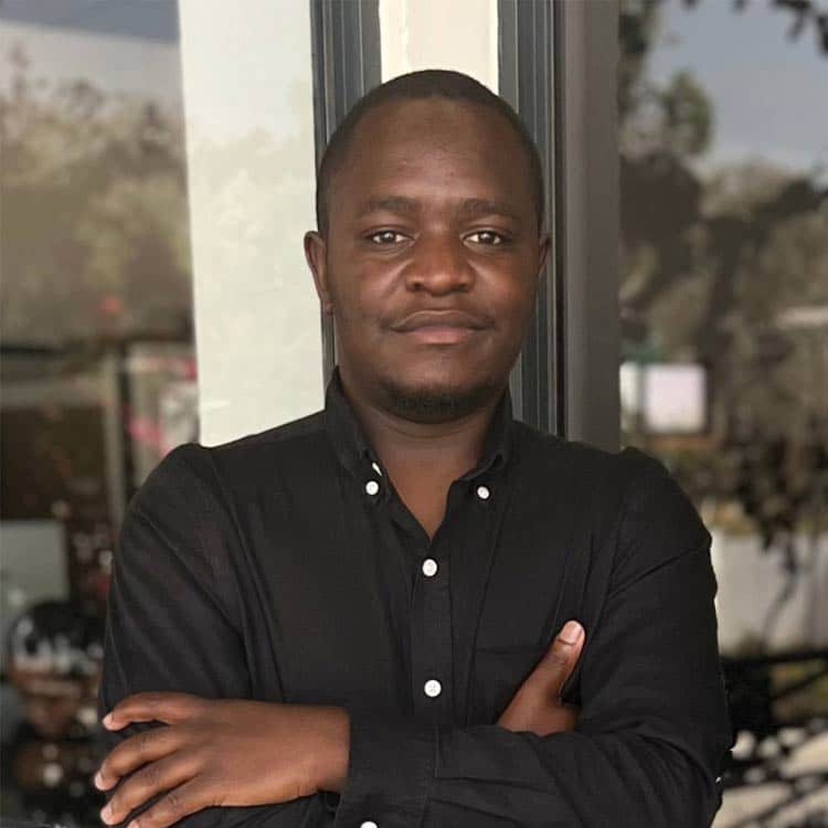 Abel Kutadza, Distribution Officer at ARC Power, wearing a black button-down shirt, standing with arms crossed in front of a glass door.