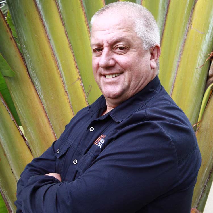 Joaquim Magalhaes, Head of Operations at ARC Power, smiling with arms crossed in front of large green palm leaves, wearing a navy ARC-branded shirt.